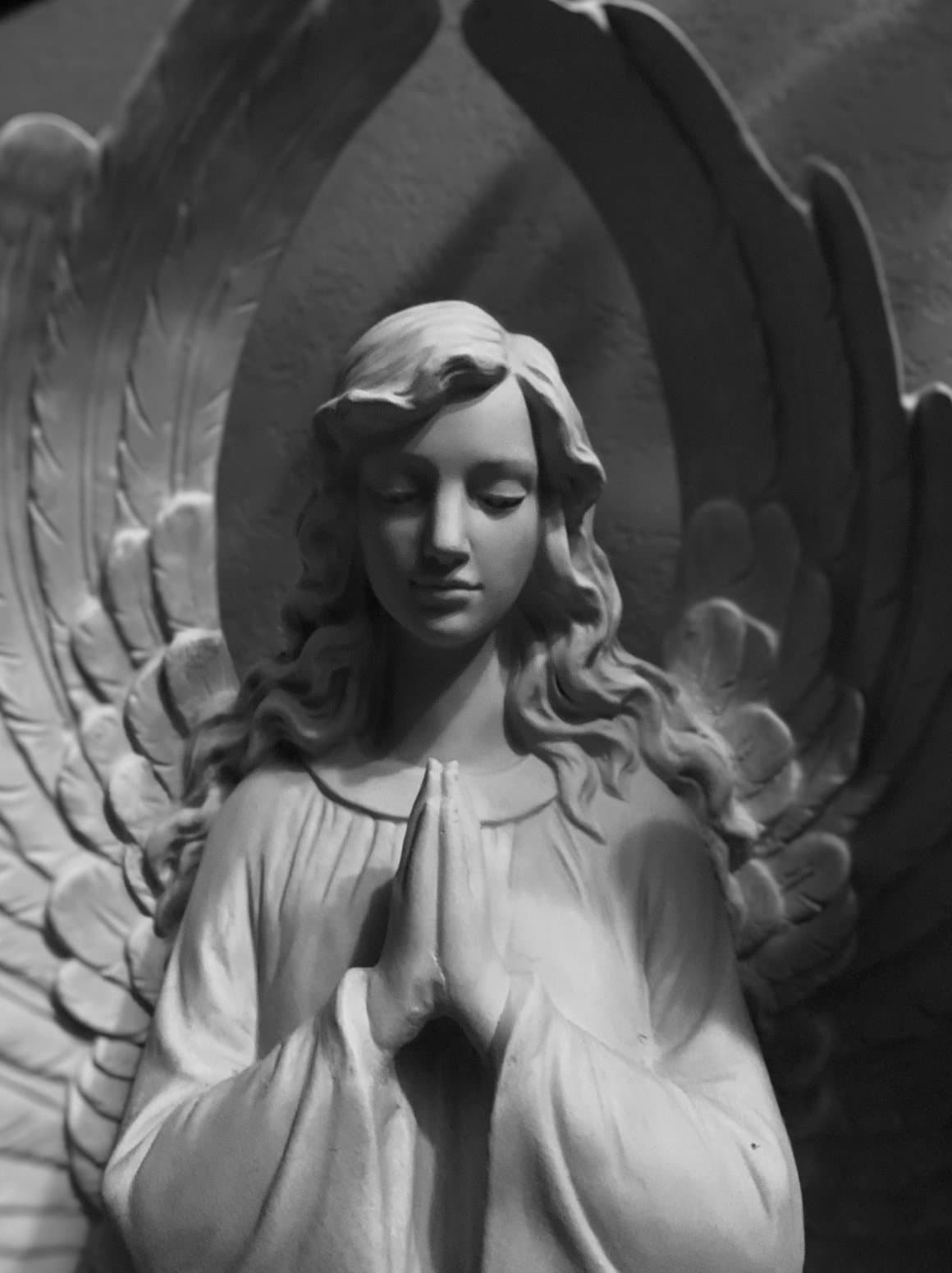 A sculpted angel with folded hands and lowered gaze, wings rising behind her.